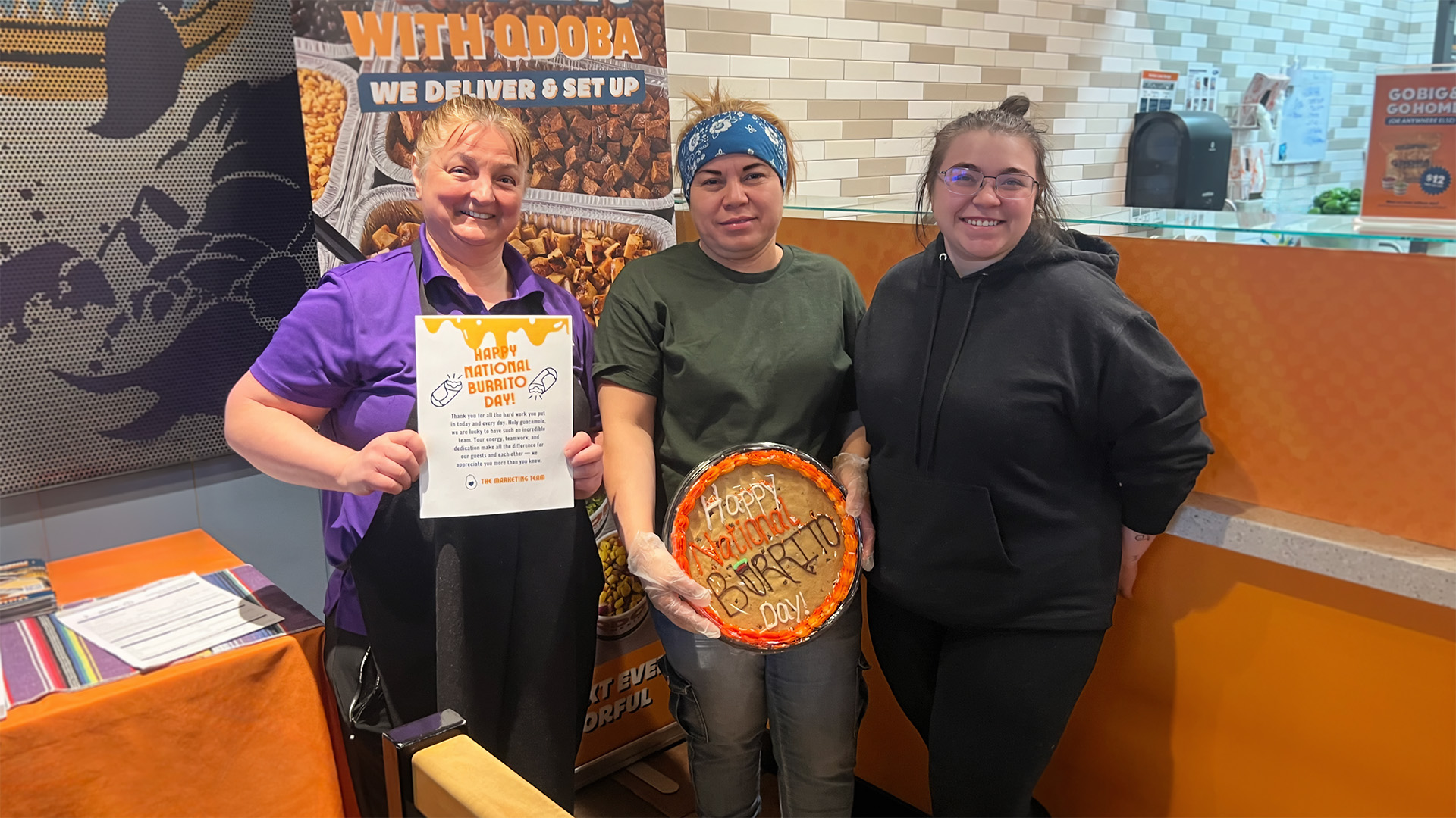 Three QDOBA team members smiling in a restaurant located in Fond du Lac, WI, holding a burrito-themed cookie cake and a “Happy National Burrito Day” appreciation sign.