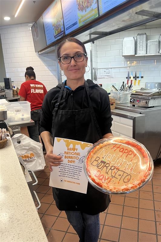 A QDOBA manager holding a decorated note and burrito-themed cookie cake, a gift for her restaurant team from Roaring Fork Restaurant Group.