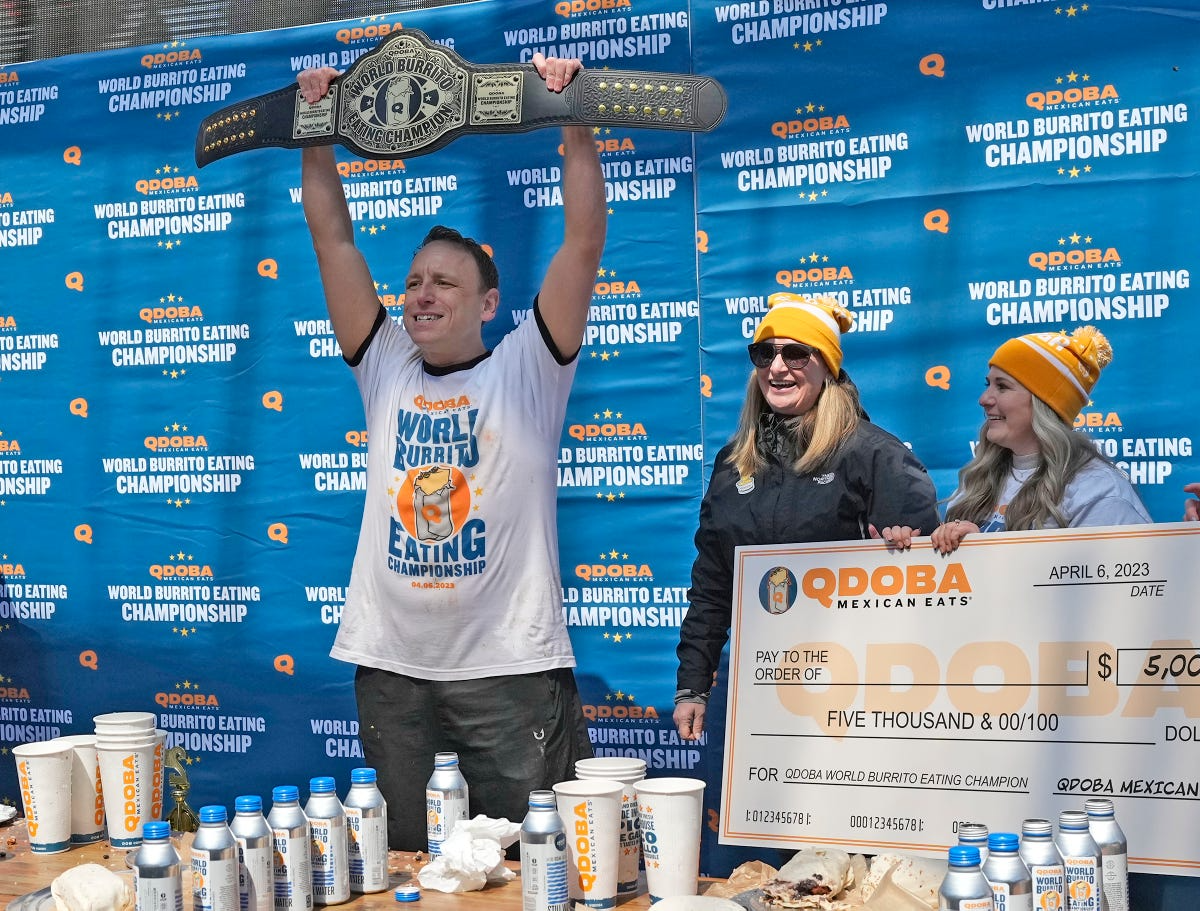 Image: Joey Chestnut, winner of the QDOBA World Burrito Eating Championship, triumphantly hoists the championship belt and proudly displays the jumbo check from QDOBA. This iconic moment captures his record-breaking feat of devouring 14.5 burritos in 10 minutes at the inaugural National Burrito Day event in Milwaukee, WI, in April 2023.