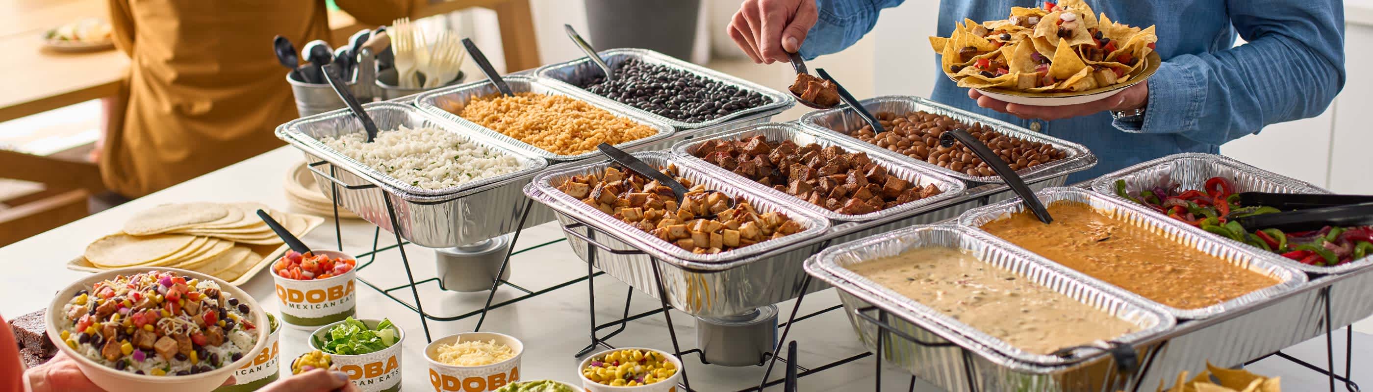 A QDOBA Catering Customizable Hot Bar with chaffing racks and serving dishes set up on a table. The hot bar includes Adobo Chicken, Grilled Steak, rice, beans, Three-Cheese Queso, spicy Queso Diablo, fajita veggies, taco tortillas, and a variety of salsas and flavorful toppings. On the far side of the table, a person is fixing a plate of nachos from the hot bar ingredients. In the bottom left side of the photo, a hand is holding a burrito bowl filled with hot bar ingredients. QDOBA Catering has made the erfect spread for a game day watch party.