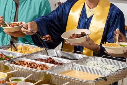 Graduation party guests build customizable QDOBA meals from a catering hot bar while celebrating a graduate in cap and gown at a graduation open house.