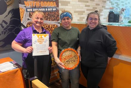 Three QDOBA team members smiling in a restaurant located in Fond du Lac, WI, holding a burrito-themed cookie cake and a “Happy National Burrito Day” appreciation sign.